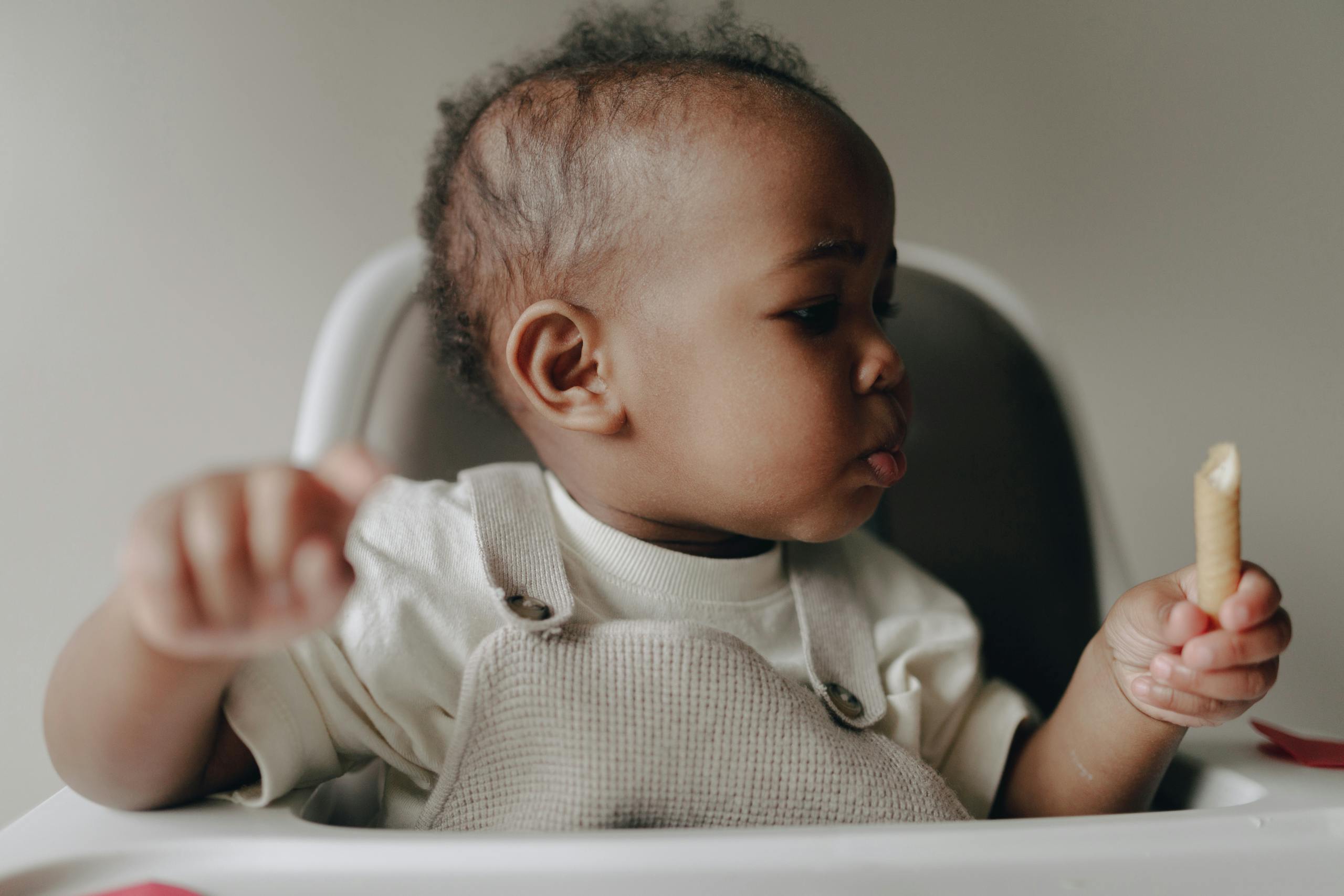 Cute baby sitting in high chair, enjoying a biscuit, with a curious facial expression.