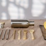 Stainless steel water bottle with a bamboo and steel cap beside steel straws, bamboo cutlery and lemons