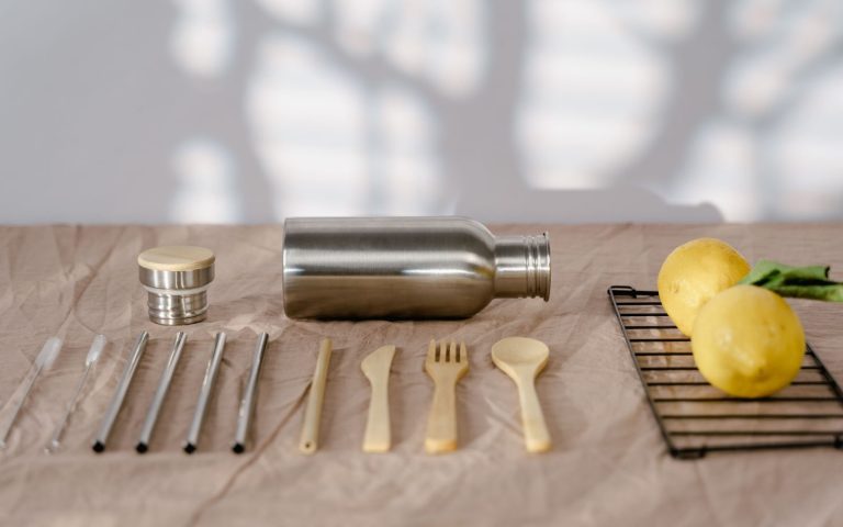Stainless steel water bottle with a bamboo and steel cap beside steel straws, bamboo cutlery and lemons