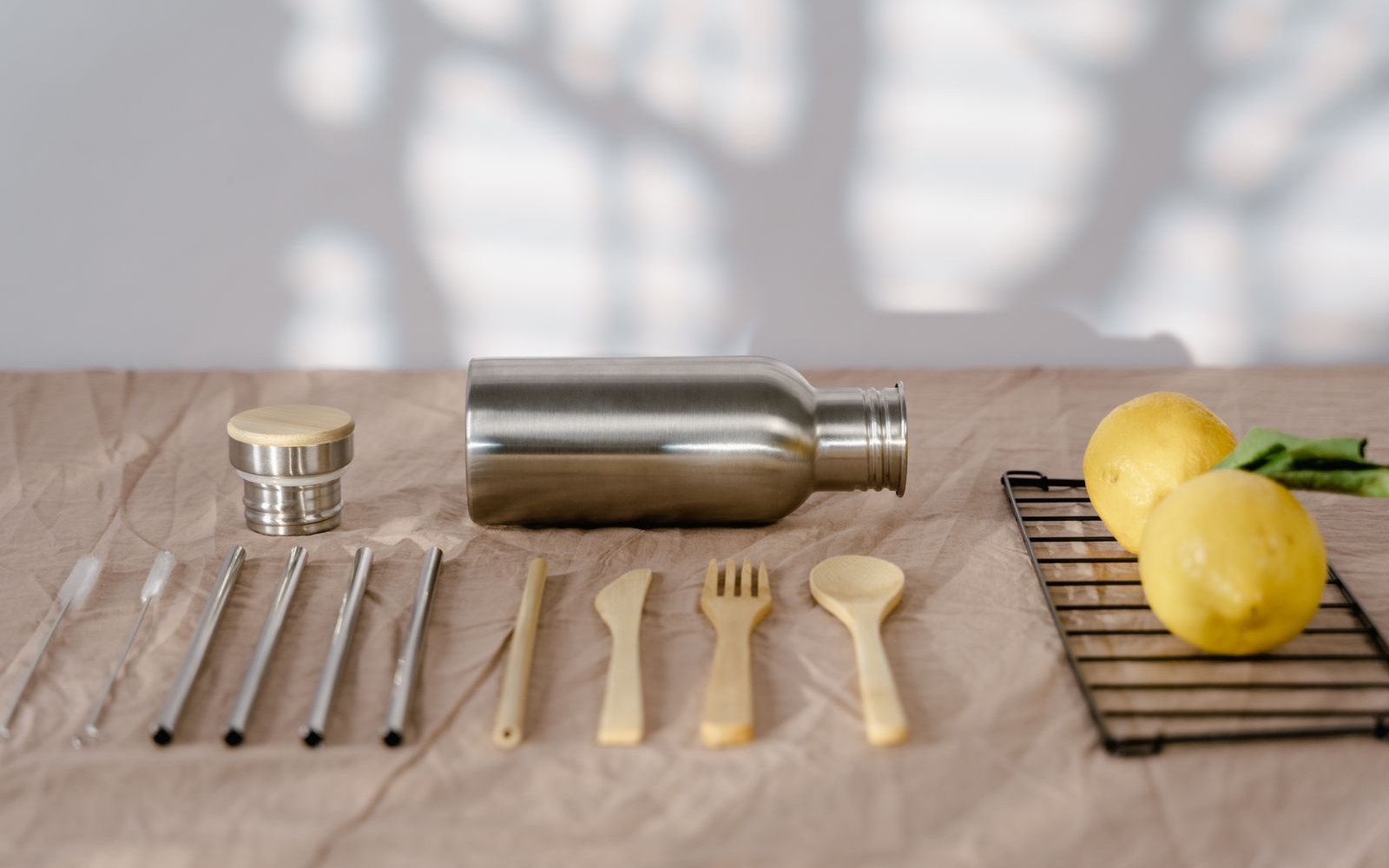 Stainless steel water bottle with a bamboo and steel cap beside steel straws, bamboo cutlery and lemons